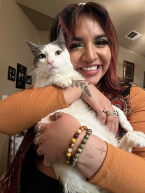 A young woman smiles happily while hugging to her chest a white cat with gray ears. She's wearing a long-sleeve orange shirt, and various bracelets, rings, and tattoos are visible on her hands and wrists.