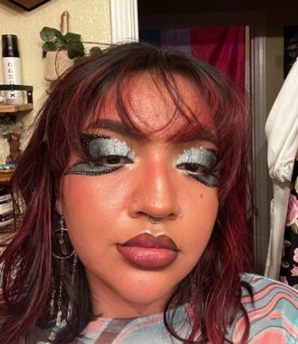 A close-up of a young woman's face to highlight the creative application of some facial makeup. Her eyes have silver eye shadow with black outlines, and her lips have a wine-colored red lipstick with black outlines. Her black hair has red highlights to match the tone of the makeup. 