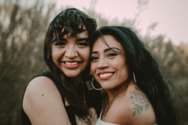 A younger and older woman stand in profile, with their faces turned toward the camera. The younger has dark hair and a big smile; the older, who's somewhat shorter, has long thick hair, a big smile, and a tattoo on one shoulder. They are outdoors with a stand of trees behind them.