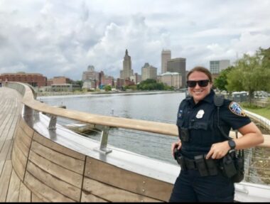 A woman, smiling, is next to a pedestrian bridge over a waterway. She is wearing a black police uniform. 