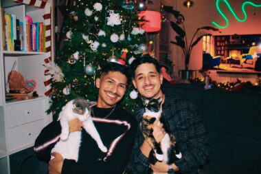 Two men sit close together on a couch in the living room of a home, each of them holding a different cat. Christmas decorations hang in the background. 