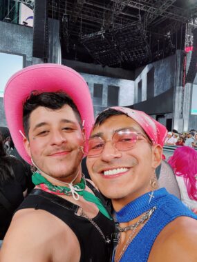 Two men embrace and smile broadly in a selfie at a concert venue. The man on the left wears a bright pink cowboy and, and the man on the right wears a pink bandana. The man on the right is also wearing matching pink sunglasses. 