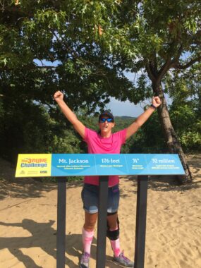 A woman stands behind a sign for Mt. Jackson with her arms raised triumphantly in the air. The sign gives more information about the mountain, which looks to be part of the "3 Dune Challenge" at the Indiana Dunes State Park. She's wearing a hat, sunglasses, a bright pink shirt, shorts, and matching pink knee-high socks. The ground is sandy and there are mature trees behind her.