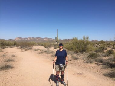 A woman stops in the middle of a trail for a photo. She's wearing a hat, sunglasses, and hiking gear, and is carrying trekking poles. She's in the middle of a desert, with a tall cactus behind her and mountains in the distance. It's a sunny, cloudless day.