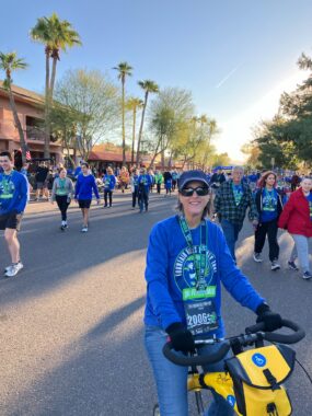 A woman seated on a walking bike poses on the side of a road for a photo. The road, likely closed off to vehicular traffic, is full of people walking and wearing blue T-shirts as part of a Turkey Trot. The woman has on the same blue shirt, along with a blue cap, sunglasses, and a lanyard with her race ID number. 