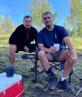 A man sits in a camp chair holding a cup while another man squats behind him. They are surrounded by green trees and grass, with a cooler at their side. 