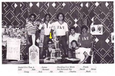 A black-and-white image, taken from a college newspaper, shows seven young men surrounded by posters and awards from their fraternity. Several are wearing T-shirts with Greek letters on them.