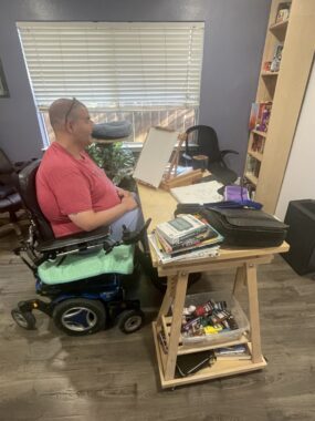 A man in a power wheelchair is shown working at a desk in his home. 