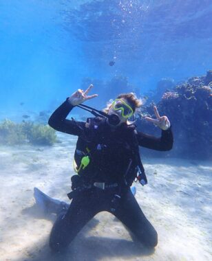 A woman flashes a victory sign as she kneels in scuba gear on the sea floor.