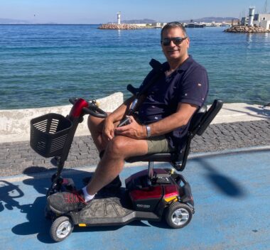 A man is shown sitting in a scooter with a beautiful view of a water and a lighthouse in the background. 