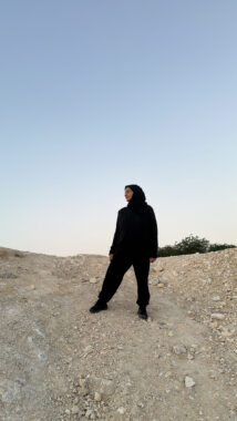 A woman wearing all black stands on a rocky path and looks off into the distance. Not much is visible around her, but it appears to be either sunrise or sunset, given the lighting.