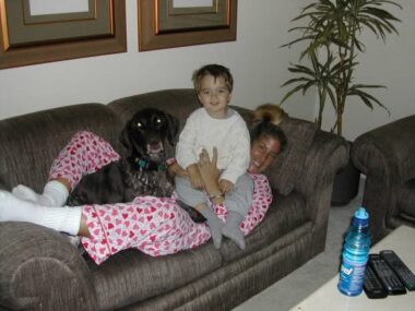 A 3-year-old boy sits on the chest of a young woman dressed in red and white pajamas with hearts all over them, as she lies on a couch. A big dog is also sitting on top of her legs. 