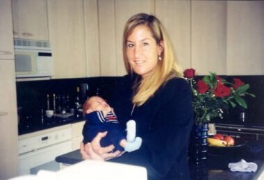 A young woman stands in a kitchen holding a newborn baby boy. A vase of roses sits on the counter behind her. 