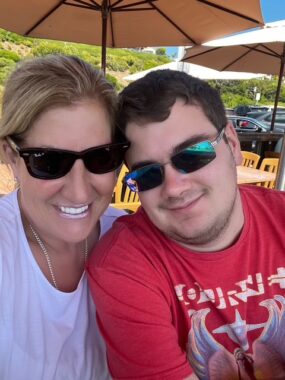 A woman smiles broadly and embraces a smiling young man next to her while both sit underneath umbrellas on a very sunny day. Both are wearing sunglasses. 