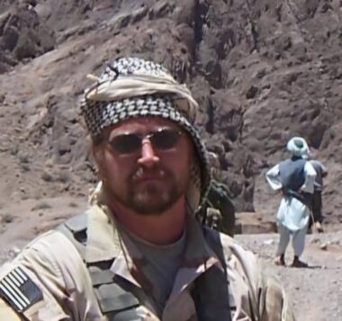 A man with a beard, mustache, shemagh, sunglasses, and U.S. military uniform faces the camera. Rocky terrain and another man in a shemagh are in the background.