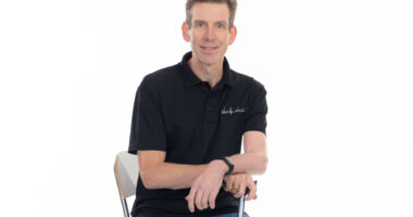 A man in a black shirt and jeans poses for a photo while seated in a chair. His hands are resting on a cane in front of him.