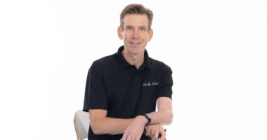 A man in a black shirt and jeans poses for a photo while seated in a chair. His hands are resting on a cane in front of him.