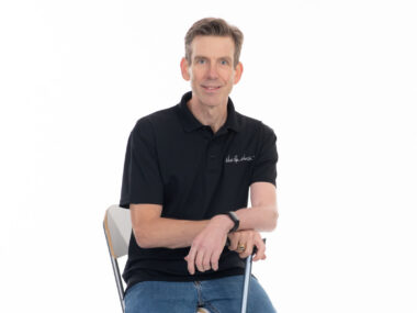 A man in a black shirt and jeans poses for a photo while seated in a chair. His hands are resting on a cane in front of him.