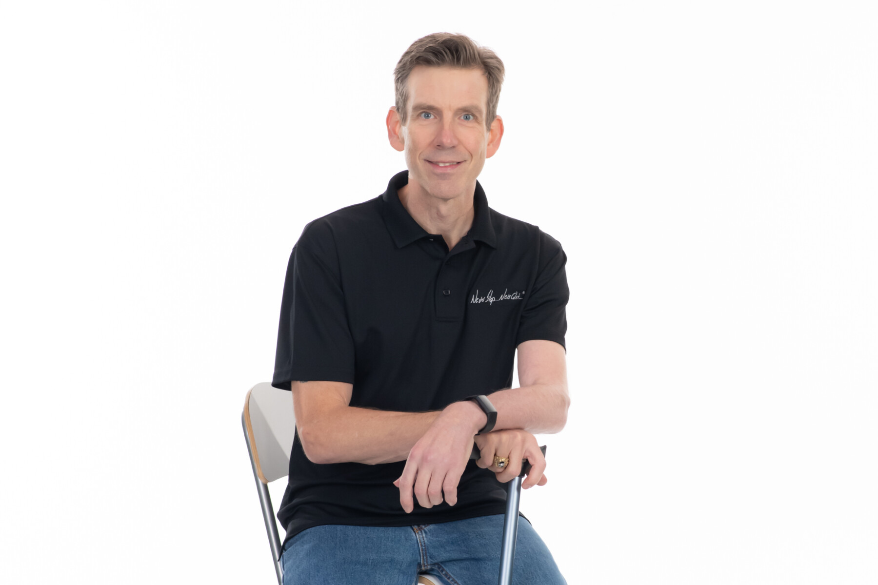 A man in a black shirt and jeans poses for a photo while seated in a chair. His hands are resting on a cane in front of him.