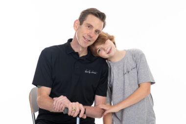 A man in a black collared shirt sits on a chair with his hands resting on a cane in front of him. His daughter stands beside him with her head tilted and resting on his shoulder. She has short red hair and is wearing a gray T-shirt.