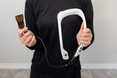 A man wearing all black holds up a PoNS device. The photo is cropped so we only see his hands and torso.