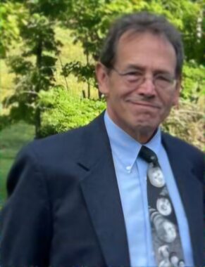 A man in a blue suit jacket and patterned tie smiles for a photo outside. There are trees behind him, and while he seems to be standing in the shade, it looks to be a sunny day.