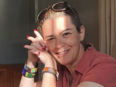 A sunny profile of a woman sitting with her elbows resting on a table and her hands clasped next to her cheek. She has sunglasses on the top of her head, and the reflection of the sunlight creates an energetic, appealing look.