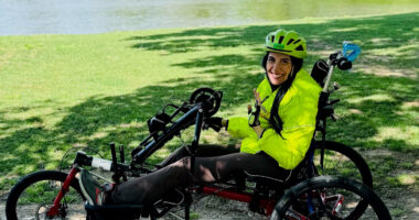 A woman wearing a bike helmet waves from the seat of her handcycle on a park pathway.