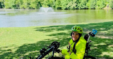 A woman wearing a bike helmet waves from the seat of her handcycle on a park pathway.