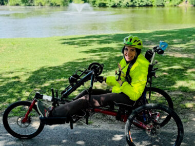 A woman wearing a bike helmet waves from the seat of her handcycle on a park pathway.