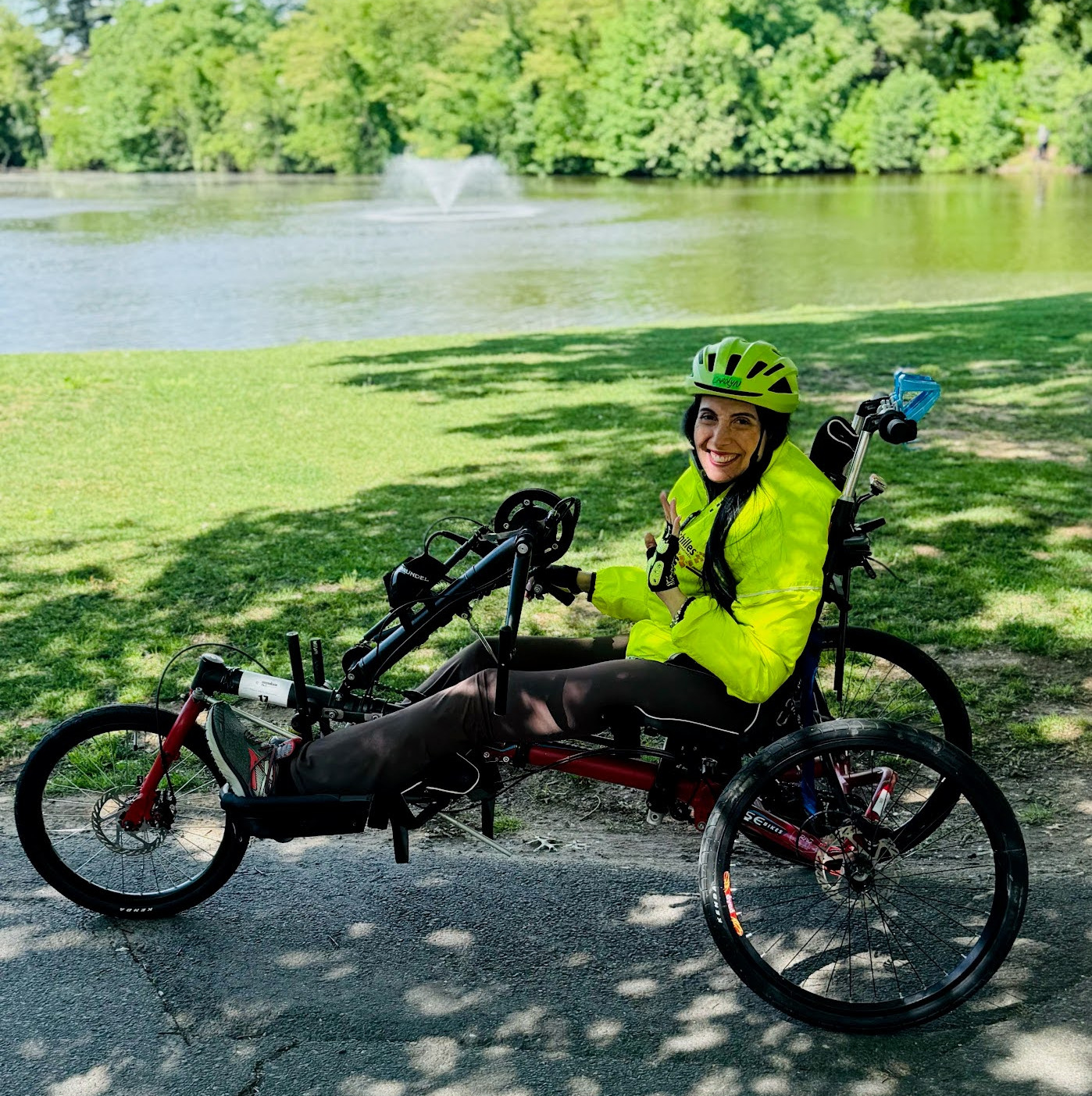 A woman wearing a bike helmet waves from the seat of her handcycle on a park pathway.