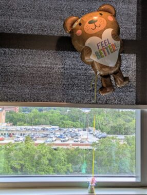 A floating balloon in the shape of a brown bear holds a heart with the words "Feel Better!" in front of a dark window screen. Below it we see many green trees surrounding a parking lot.