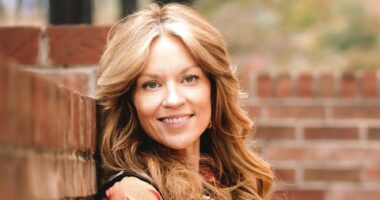 A woman with long flowing hair stands in front of the corner of two adjoining low brick walls. The photo projects brightness, as if it's summertime, and her reddish/blond hair blends naturally with the color of the bricks in the wall.