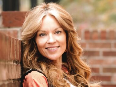 A woman with long flowing hair stands in front of the corner of two adjoining low brick walls. The photo projects brightness, as if it's summertime, and her reddish/blond hair blends naturally with the color of the bricks in the wall.