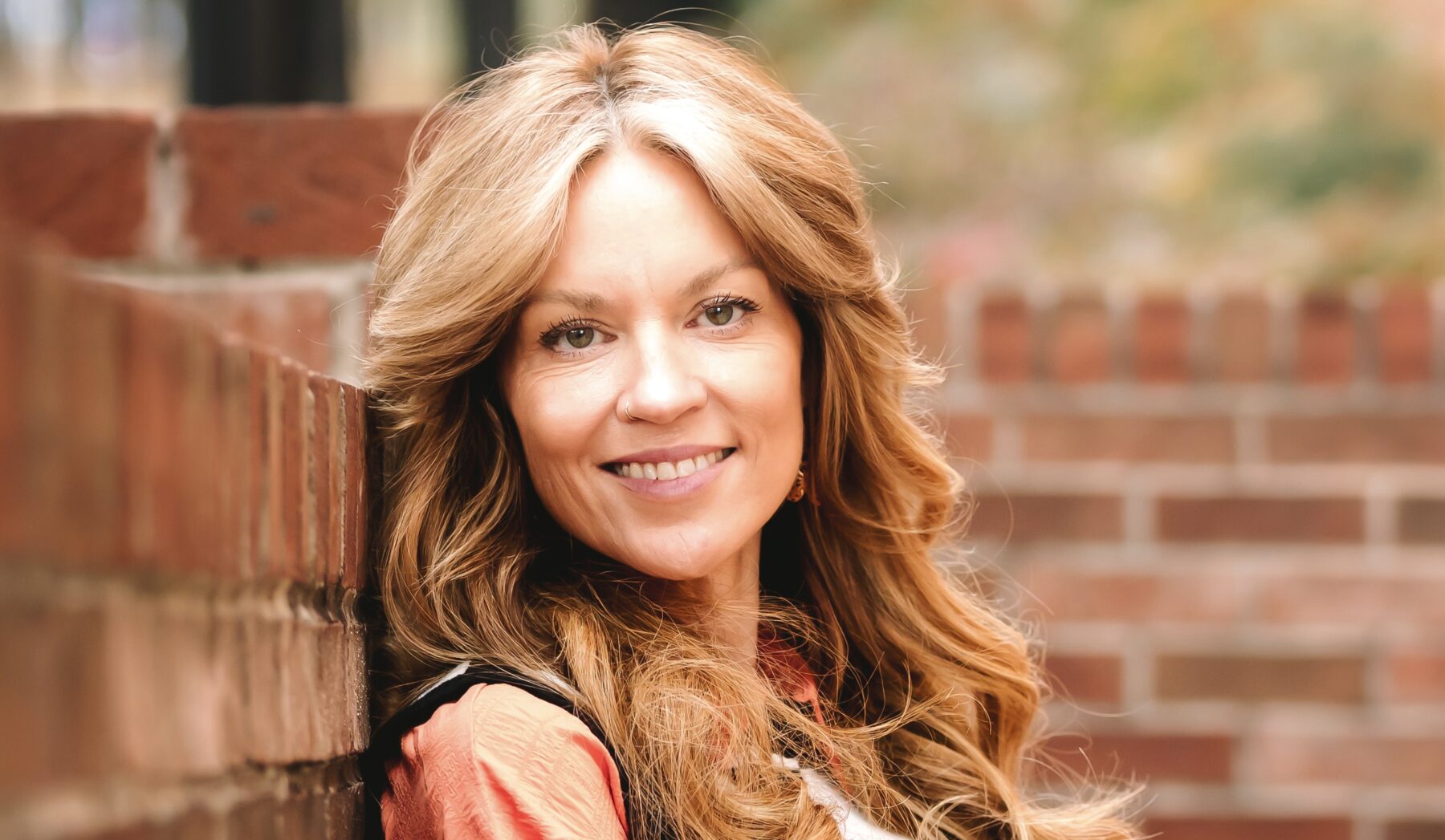 A woman with long flowing hair stands in front of the corner of two adjoining low brick walls. The photo projects brightness, as if it's summertime, and her reddish/blond hair blends naturally with the color of the bricks in the wall.