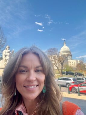 A woman takes a selfie of her face as she stands in front of the Capitol building in Washington, D.C., which can be seen over her left shoulder. It's a sunny, cloudless day. Cars are parked between her and the Capitol, giving us a sense of how far away she is from it, probably about a 5- to 10-minute walk.