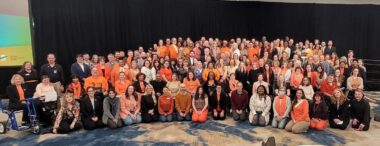 A horizontal photo shows a large group of people, most of them wearing orange T-shirts, the color symbolizing MS awareness. It's hard to tell, but it seems like there are 70-100 people in the photo.