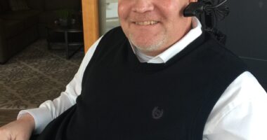 A side profile of a man in a long-sleeved white dress shirt with a black vast over it. He's sitting down and smiling as he looks at the camera to his left. He has a head piece with headphones and a microphone.