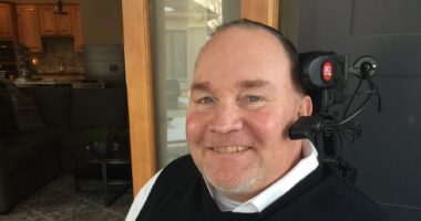 A side profile of a man in a long-sleeved white dress shirt with a black vast over it. He's sitting down and smiling as he looks at the camera to his left. He has a head piece with headphones and a microphone.