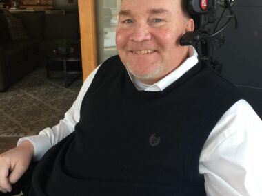 A side profile of a man in a long-sleeved white dress shirt with a black vast over it. He's sitting down and smiling as he looks at the camera to his left. He has a head piece with headphones and a microphone.