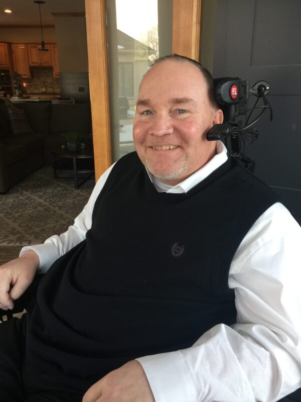 A side profile of a man in a long-sleeved white dress shirt with a black vast over it. He's sitting down and smiling as he looks at the camera to his left. He has a head piece with headphones and a microphone.