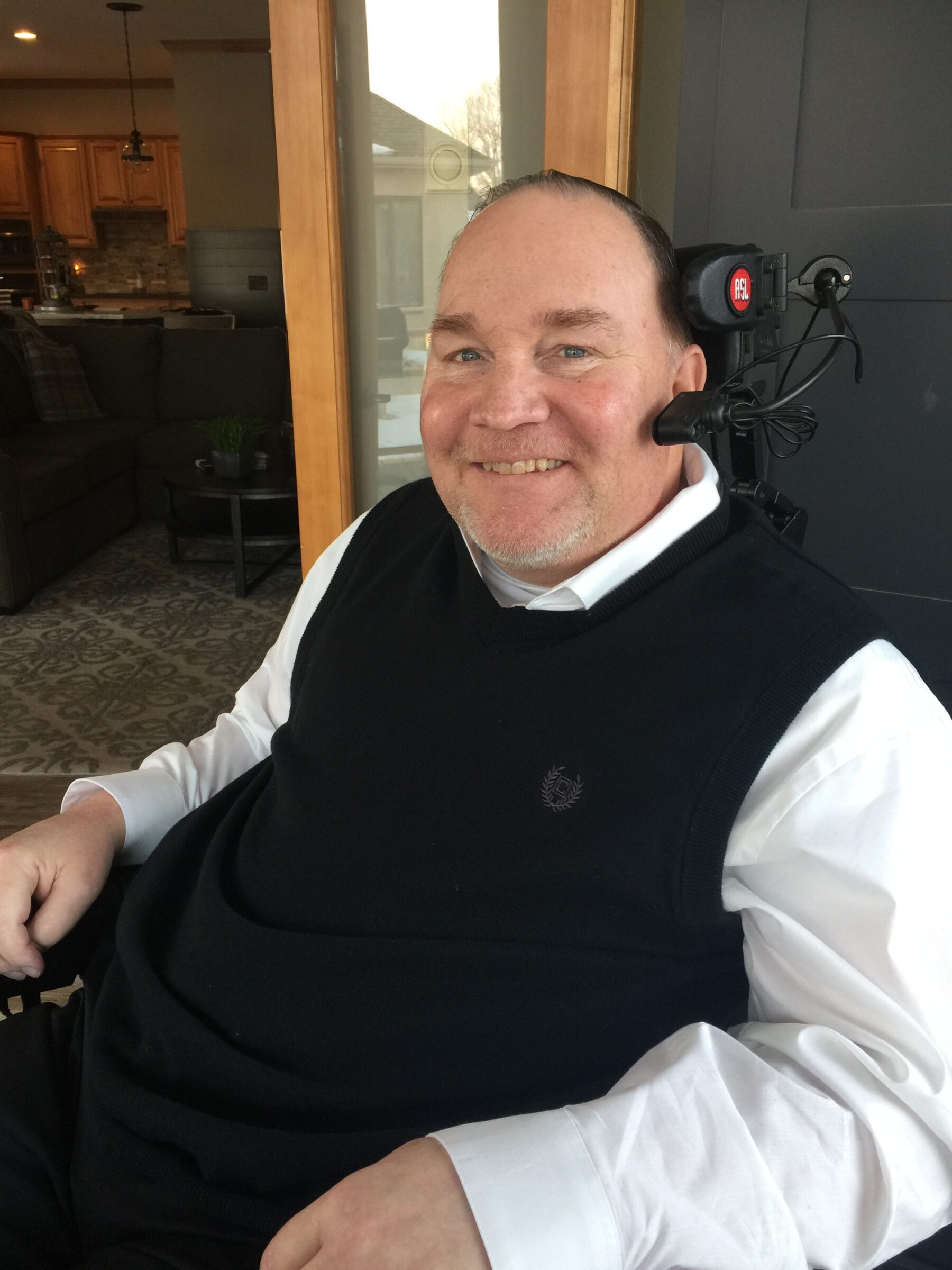 A side profile of a man in a long-sleeved white dress shirt with a black vast over it. He's sitting down and smiling as he looks at the camera to his left. He has a head piece with headphones and a microphone.