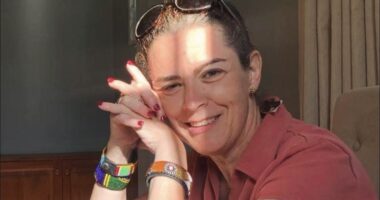 A close-up of a woman in a red shirt smiling and looking at the camera, with the right side of her head resting slightly and gently on her folded hands. Sunshine and shadows reflect off her face.