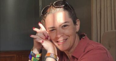 A close-up of a woman in a red shirt smiling and looking at the camera, with the right side of her head resting slightly and gently on her folded hands. Sunshine and shadows reflect off her face.