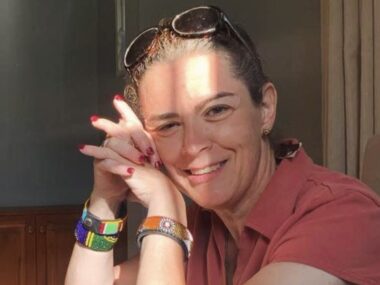 A close-up of a woman in a red shirt smiling and looking at the camera, with the right side of her head resting slightly and gently on her folded hands. Sunshine and shadows reflect off her face.