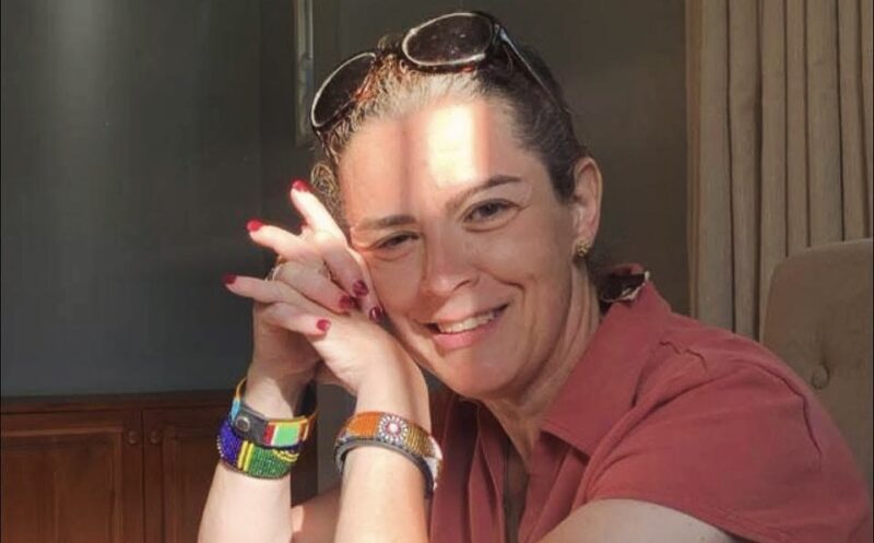 A close-up of a woman in a red shirt smiling and looking at the camera, with the right side of her head resting slightly and gently on her folded hands. Sunshine and shadows reflect off her face.