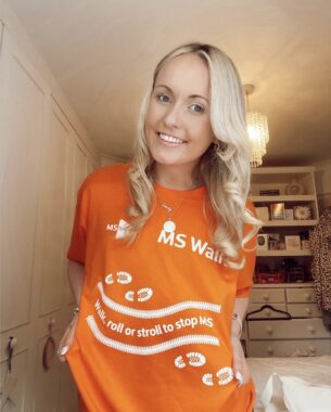 A young woman smiles for a photo while wearing an orange T-shirt that reads "MS Walk: Walk, roll, or stroll to stop MS." She appears to be in her bedroom.