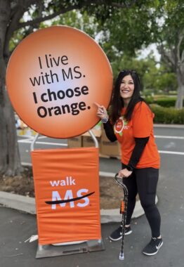 A woman with a bright, orange T-shirt smiles while pointing to a large orange display that says, "I live with MS. I choose orange." Lower on it, it says, "Walk MS." She's on a paved path in a tree-lined outdoor area and balances her left hand on a cane.
