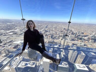 A woman sits in a clear observation box at the top of a skyscraper in Chicago. The glass walls and floor offer a sweeping view of the city.