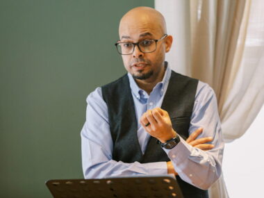 A bald man in a dress shirt and vest, and with glasses, stands at a podium delivering a sermon.